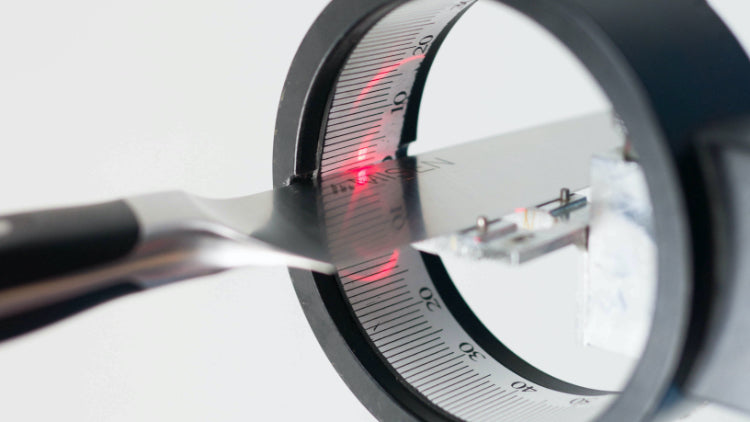 Metal knife tip near a circular measuring ring with numbered scale, red laser dot on the blade and LED circuit inside.