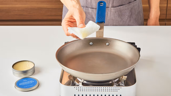 Hand wiping interior of metal frying pan with paper towel on a portable gas burner; blue handle and open tin nearby.