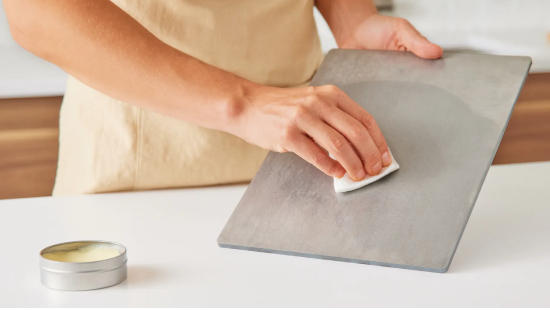 Hands wiping a rectangular metal sheet with a white pad on a counter; small round tin nearby