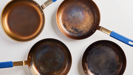 Four patinated copper-colored frying pans with blue handles arranged on white background