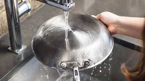 Hand holding upside-down stainless steel frying pan under running faucet, water cascading off pan into sink