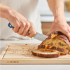 Using a blue Misen eight inch serrated knife to cut a crusty loaf of bread atop a Misen wooden cutting board