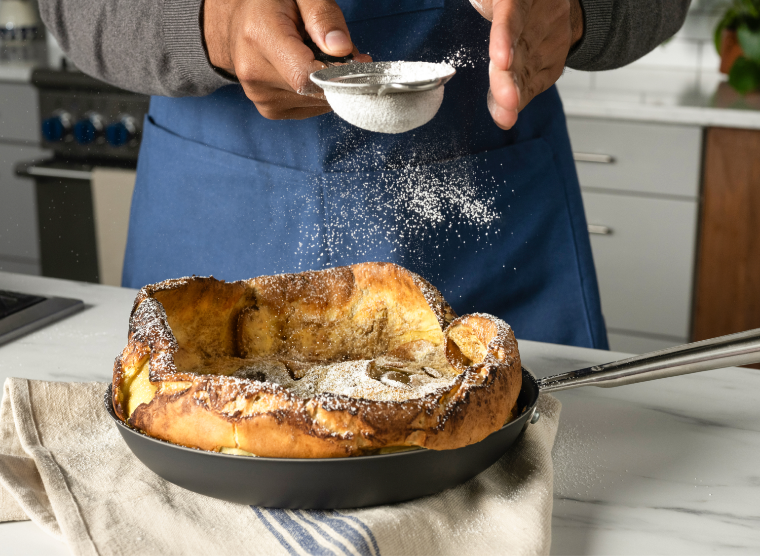 A Dutch baby pancake, golden brown and puffed, sits in a Misen Carbon Nonstick™ Frying Pan on a kitchen counter. A person, wearing a blue Misen Apron, is sifting powdered sugar over the pancake from above.