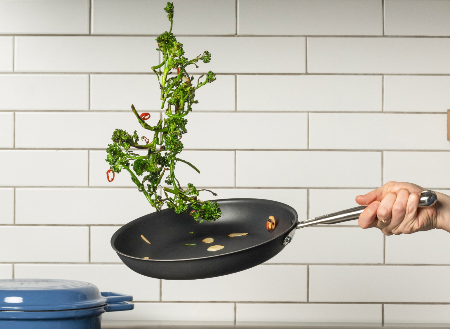 A Misen Carbon Nonstick™ Frying Pan, held by a hand, tosses broccoli and sliced garlic into the air against a white subway tile backsplash. A blue Misen Grill Dutch Oven is visible on the stove in the foreground.