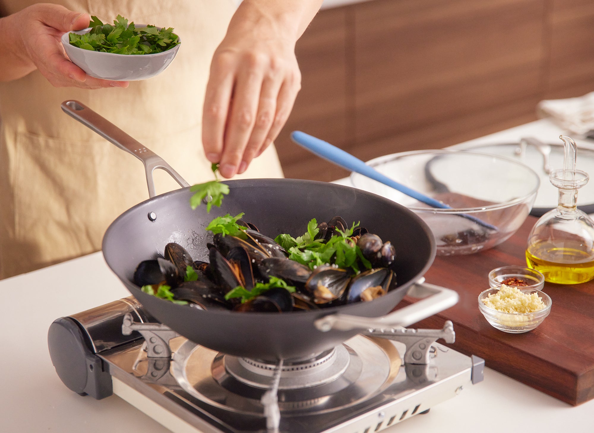{{12-inch}} A chef sprinkles herbs into a pile of muscles inside a Misen Carbon Steel wok, which sits atop a single burner. Next to the wok is a cutting board with assorted garnishes and oils.