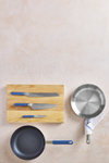 A three piece blue Misen Knife Set on top of a wooden cutting board. Next to the cutting board is an Original Misen Nonstick Pan and a Stainless Steel Pan with a Misen Kitchen Towel wrapped around its handle.