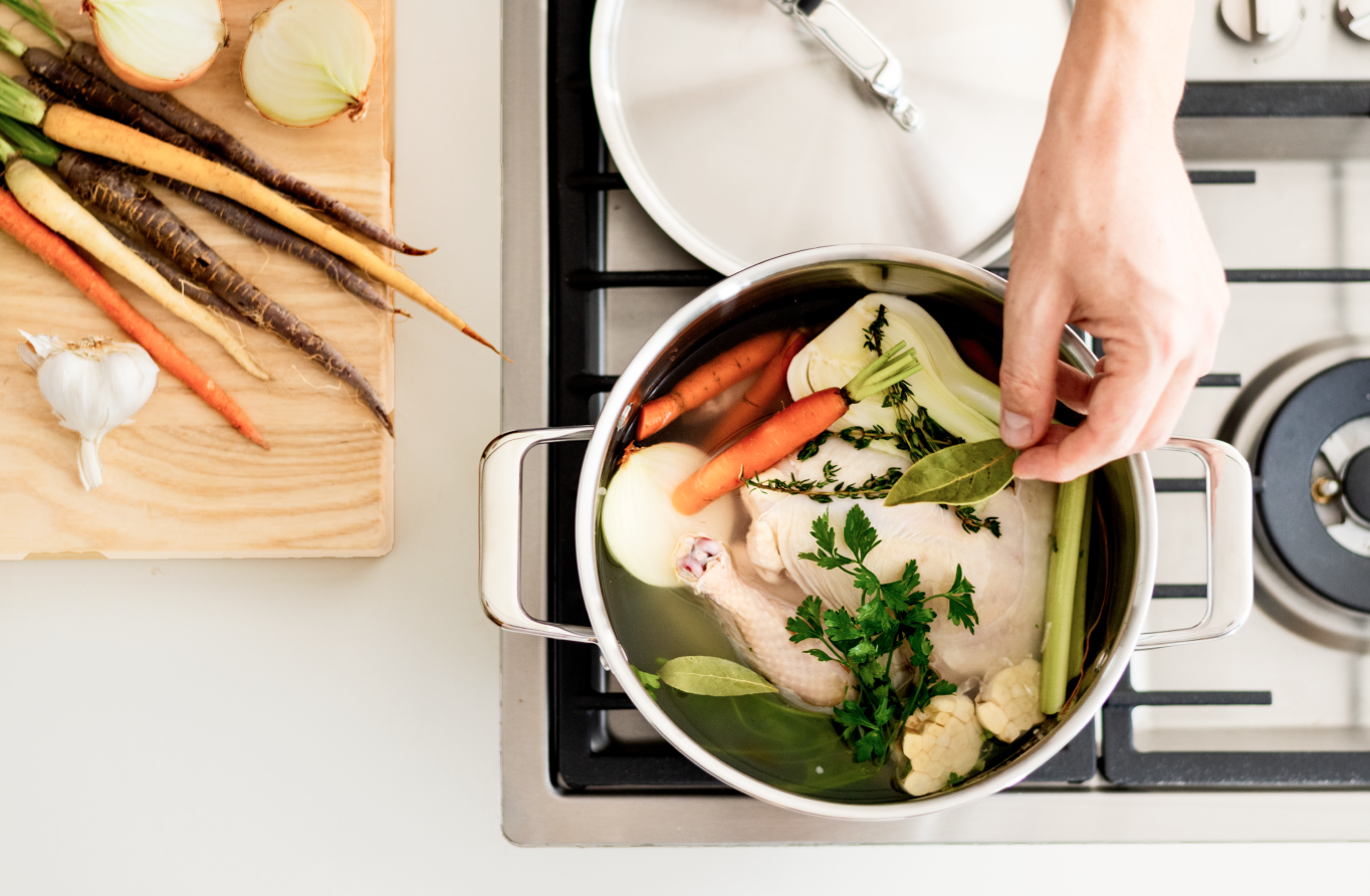 Placing a whole chicken, carrots, celery, onions, fresh parsley, and basil into an 8 quart stainless steel Misen Stockpot to make chicken soup atop a stovetop.