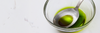 A close-up view shows a stainless steel Misen Measuring Chef Spoon resting in a clear glass bowl containing a green olive oil. The spoon is etched with measurements for 1 tbsp and 1 tsp.