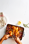 A close-up overhead shot shows a person's hands carving a roasted chicken in a rectangular roasting pan. The chicken is surrounded by roasted red onions and lemon slices. The pan has two handles, one visible in blue and the other with a silver finish. To the left, a white plate holds two pieces of silverware and a folded striped towel. In the upper right corner, a small glass bowl contains lemon slices and a sprig of thyme. The background is a white marble surface.