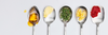 Five stainless steel chef spoons in a row, each holding a different ingredient. From left to right: a spoon with red tomato paste, a spoon with yellow bow-tie pasta, a spoon with dr thyme, a spoon with pale yellow butter, and a spoon with dark amber honey.