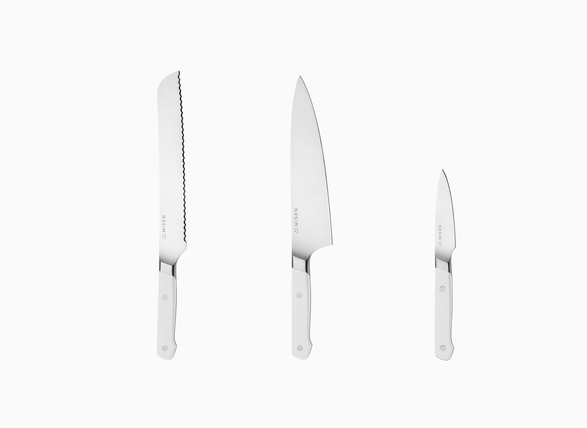 The image displays a set of three Misen knives with white handles and AUS-10 steel blades, arranged vertically against a white background. From left to right, the knives decrease in size. The leftmost knife is the longest and features an 8 inch serrated blade. The middle knife has a smooth, curved blade and is an 8 inch chef's knife. The rightmost knife is the smallest, with a straight, smooth blade, resembling a paring knife. Each knife has the word "MISEN" faintly visible near the handle on the blade.