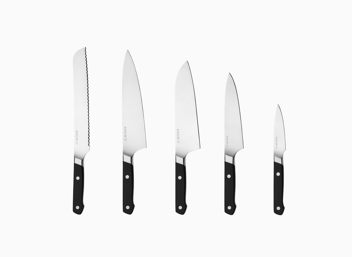 The image displays a set of five Misen knives with black handles and AUS-10 blades, arranged vertically against a white background. From left to right, the knives decrease in size. The leftmost knife has an 8" serrated blade. The second knife from the left is an 8" chef's knife. The middle knife is an 8" santoku knife. The fourth knife from the left is a 5.5" utility knife. The rightmost knife is a 3.5" paring knife. Each knife has the word "MISEN" faintly visible near the handle on the blade.