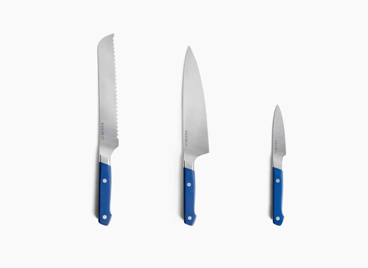 Three blue-handled knives are shown upright against a white background. From left to right, there is an 8 inch Serrated Knife 2.0, an 8n inch Chef's Knife 2.0, and a 3.5 inch Paring Knife 2.0. Each knife has a silver-toned ACUTO440 steel blade with "MISEN" and a logo engraved on it. The handles are a vibrant blue with visible rivets.