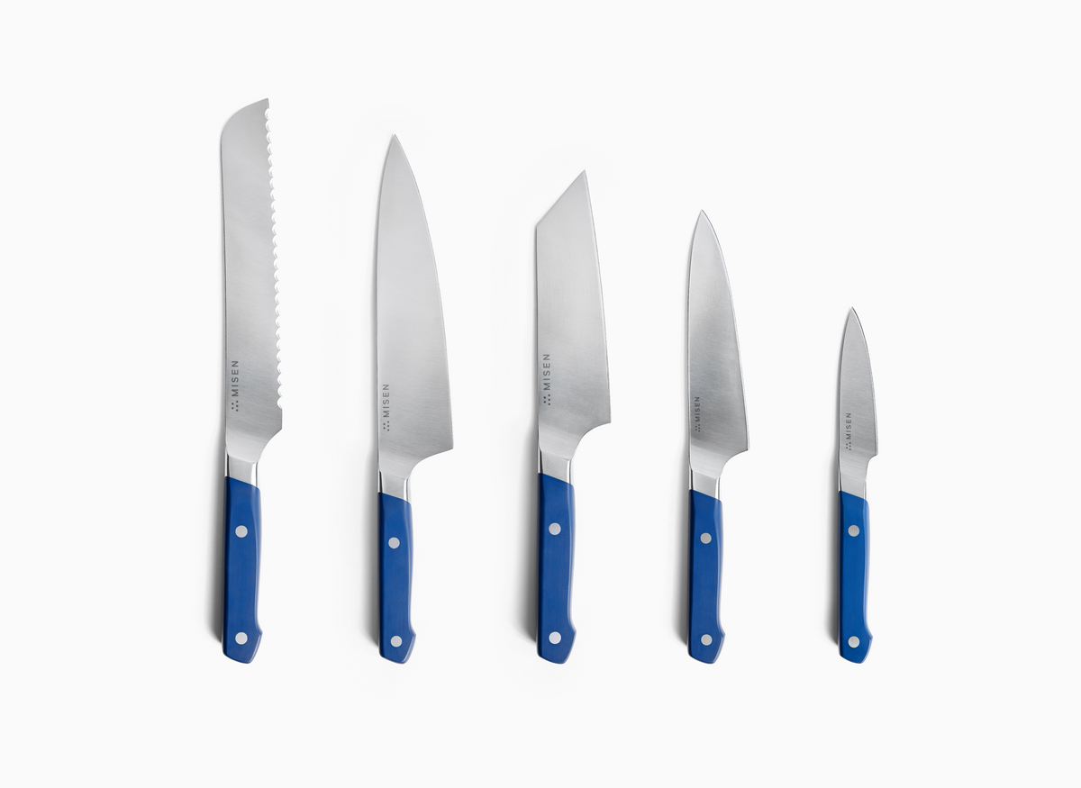 A set of five ACUTO440 steel Misen knives with blue handles are arranged in a row, from largest to smallest, on a white background. The largest 8 inch knife on the left has a serrated edge. The other four knives have smooth blades, with the second largest having a classic 8 inch chef's knife shape and the third largest, a 6 inch bunka knife, having a more angular, cleaver-like shape. The fourth knife is a 5.5 inch utility knife, and the smallest on the right is a 3.5 inch paring knife.