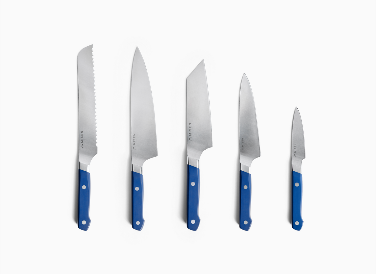 A set of five ACUTO440 steel Misen knives with blue handles are arranged in a row, from largest to smallest, on a white background. The largest 8 inch knife on the left has a serrated edge. The other four knives have smooth blades, with the second largest having a classic 8 inch chef's knife shape and the third largest, a 6 inch bunka knife, having a more angular, cleaver-like shape. The fourth knife is a 5.5 inch utility knife, and the smallest on the right is a 3.5 inch paring knife.