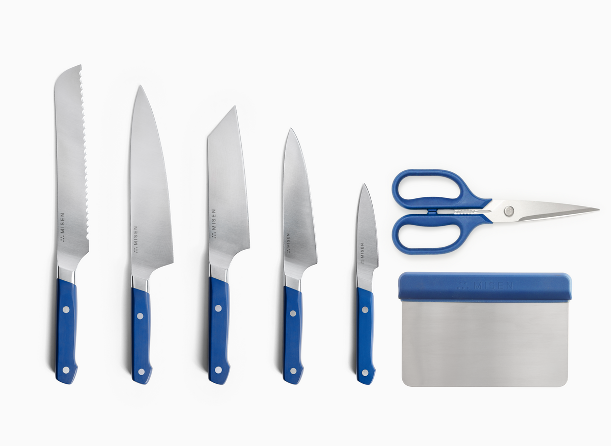 A high-angle shot on a white background shows five Misen ACUTO440 steel knives with blue handles, a pair of standard blue-handled Misen kitchen shears, and a Misen bench scraper. The knives are arranged from left to right in descending order of size: an 8 inch serrated knife, an 8 inch chef's knife, a 6 inch bunka knife, a 5.5 inch utility knife, and a 3.5 inch paring knife. To the right of the knives, the kitchen shears and bench scraper are stacked vertically.