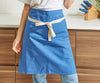 A blue Misen apron is worn on a chef and folded over to create a bistro apron. The chef is visible from the chest down and is leaning against a kitchen counter.