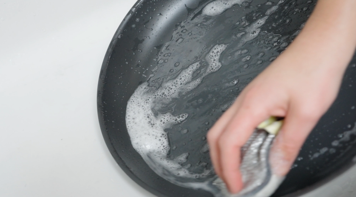 Hand scrubbing a dark nonstick frying pan with soapy sponge