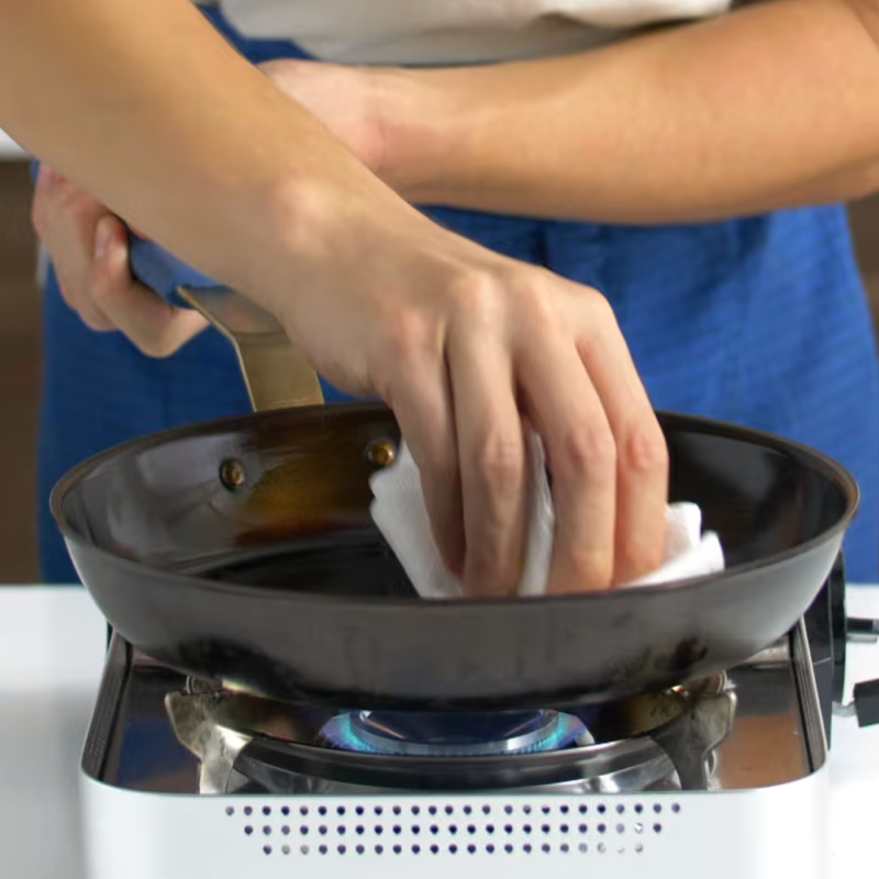 Seasoning a carbon steel pan with oil and paper towel over gas burner