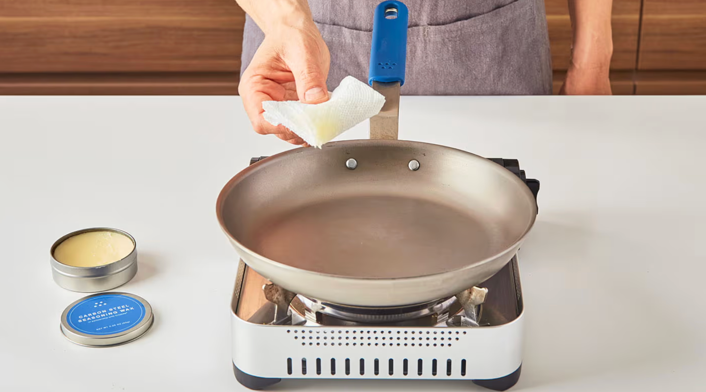 Seasoning a carbon steel pan with wax using a paper towel on a portable gas burner
