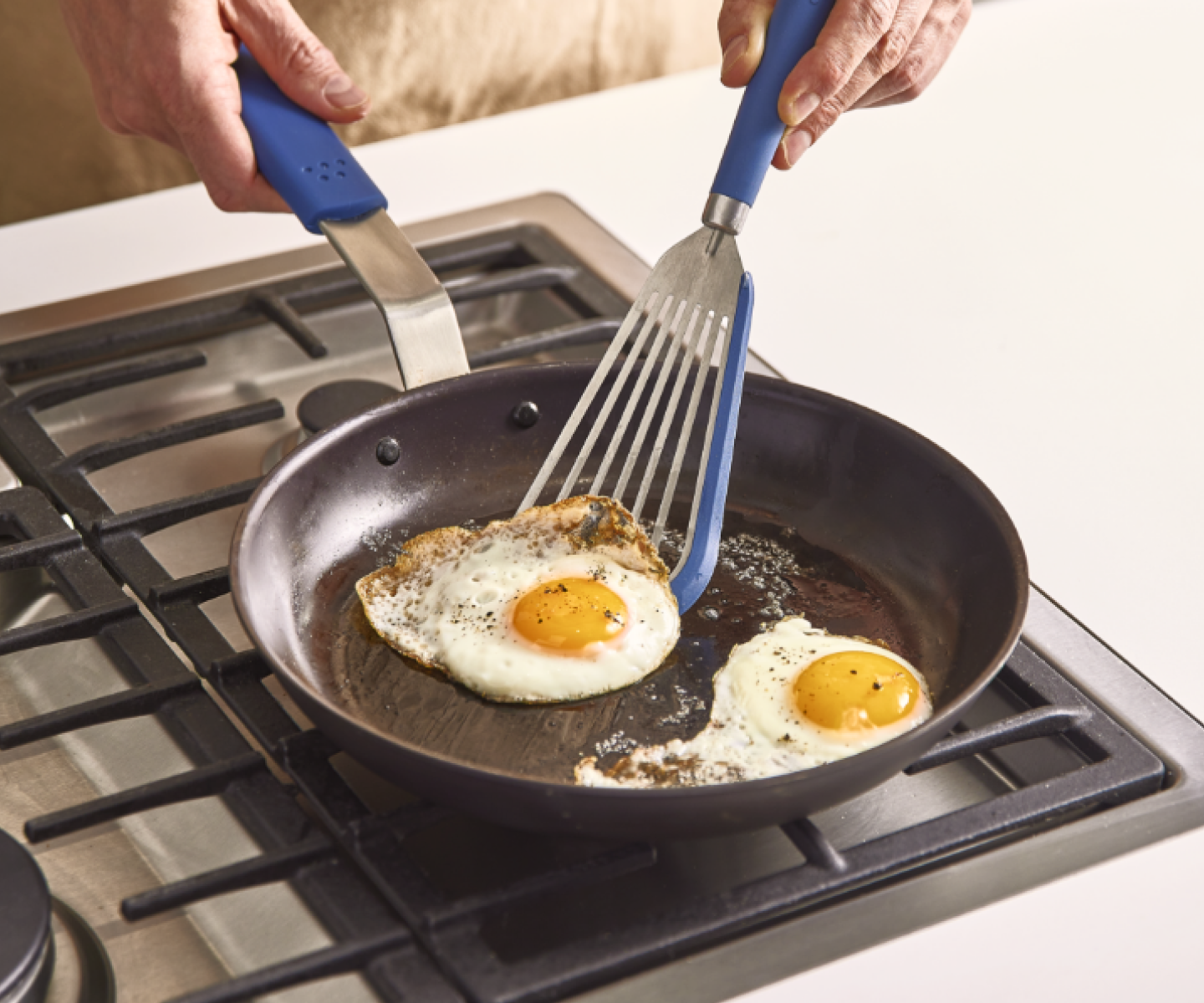Cooking sunny side up eggs in a skillet on a gas stove with blue-handled spatulas