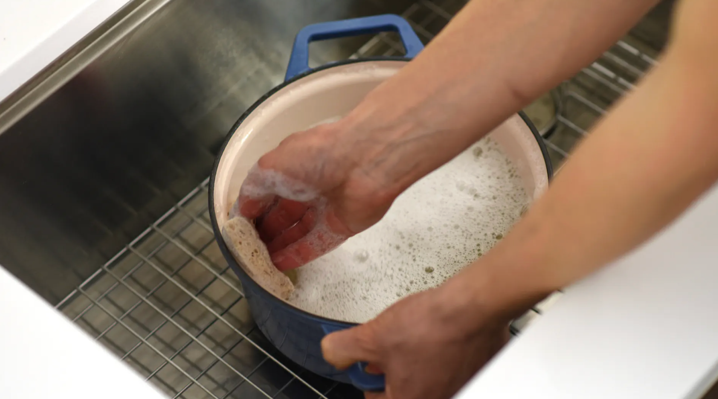 Hands scrubbing blue Dutch oven with soapy water in kitchen sink