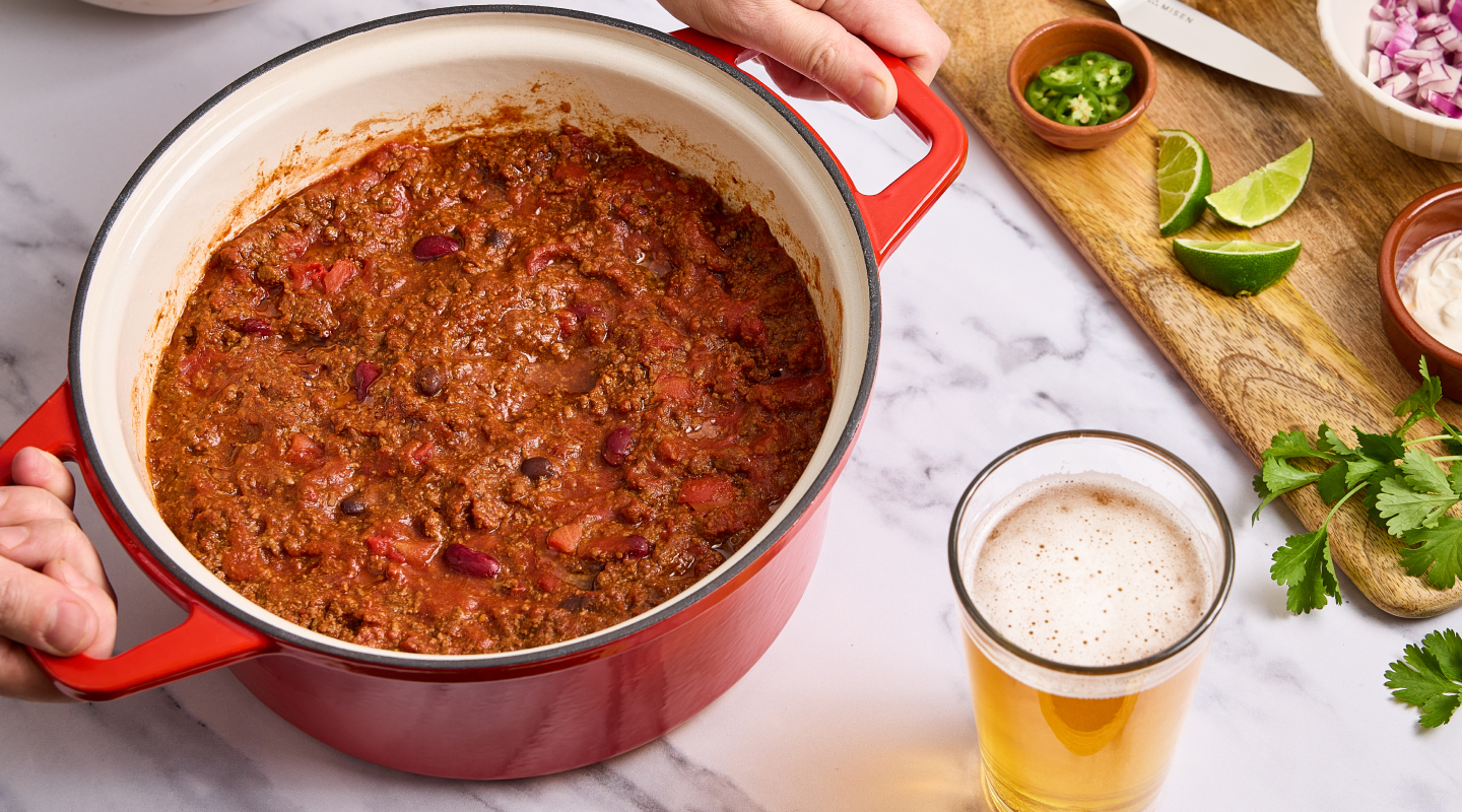 Homemade beef chili in a red Dutch oven with lime, jalapeño, red onion, cilantro, and a cold beer