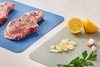 Three raw steaks rest on a large Misen Cutting Sheet, while sliced lemon, garlic, and parsley sit nearby on a small Misen Cutting Sheet.