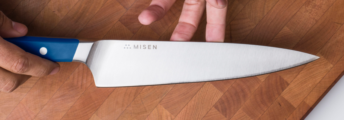 A bird’s eye view of the blade of a blue Misen Chef’s knife, in a chef’s hands, on a wood cutting board.