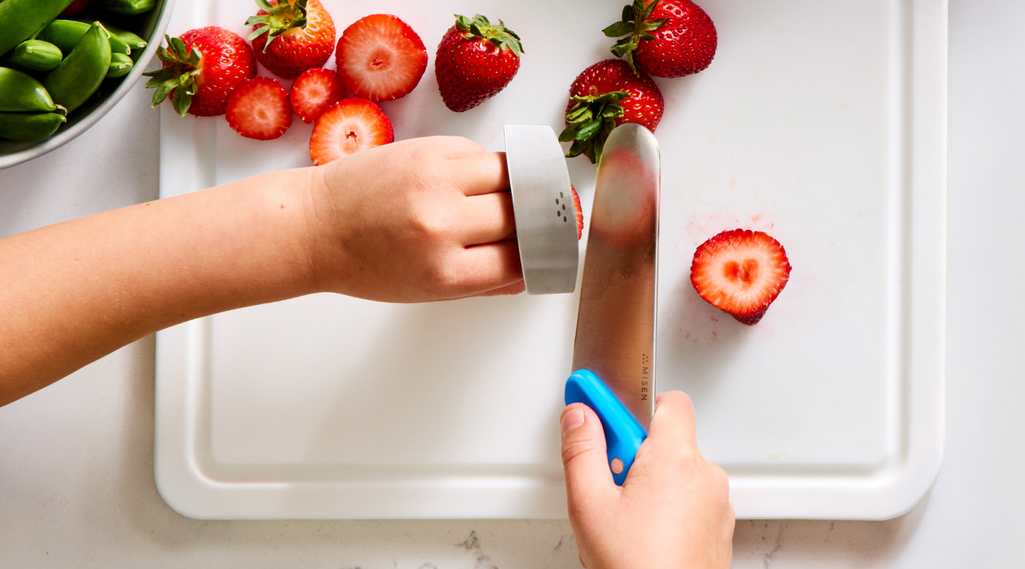 Child using Misen kids knife with finger guard to cut fresh strawberries on white cutting board