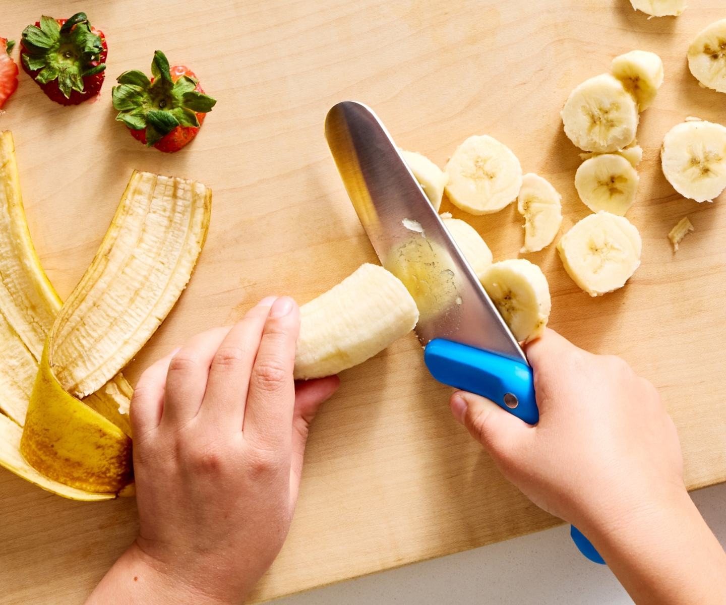 Child slicing banana on wooden cutting board with kid-safe knife next to fresh strawberries