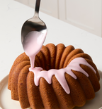 Pink glaze being spooned over a golden brown bundt cake on a white plate
