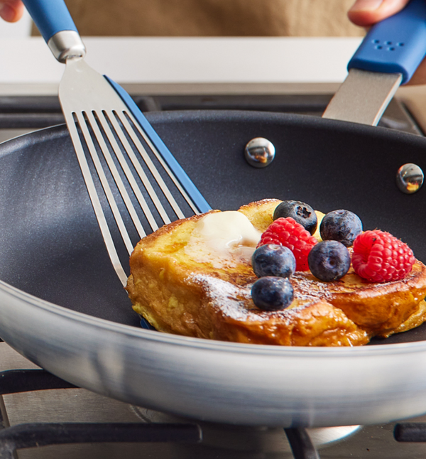 French toast topped with blueberries, raspberries, and butter cooking in a nonstick skillet with blue-handled spatula
