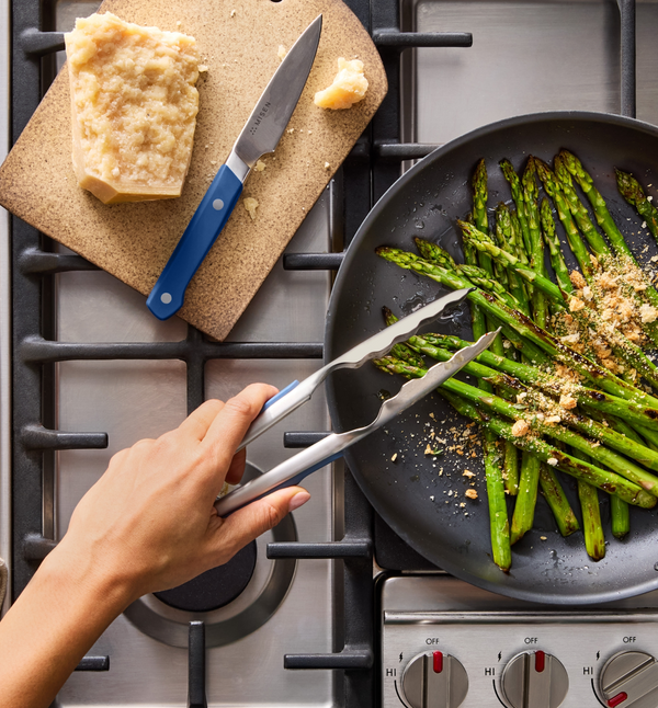 Sautéed asparagus with parmesan in a dark skillet on gas stove using Misen tongs and knife