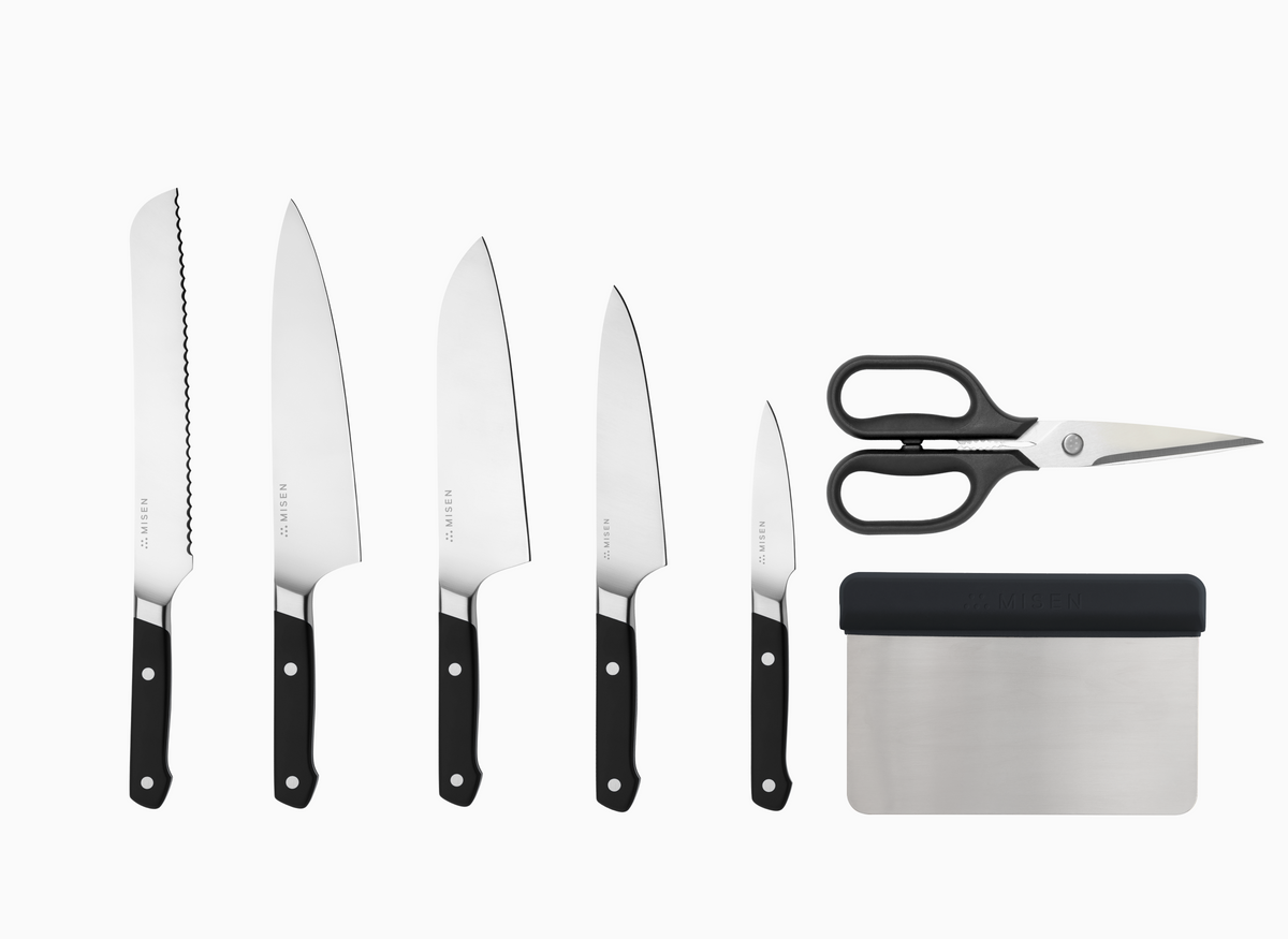 A 7 pc knife set with black handles and AUS-10 steel blades, arranged vertically against a white background. The leftmost knife has an 8" serrated blade. The 2nd knife is an 8" chef's knife. The 3rd knife is an 8" santoku knife. The 4th knife is a 5.5" utility knife. The 5th knife is a 3.5" paring knife. To the right of the knives, there is a pair of standard kitchen shears and a rectangular bench scraper, both with black handles. Each item has the word "MISEN" visible.