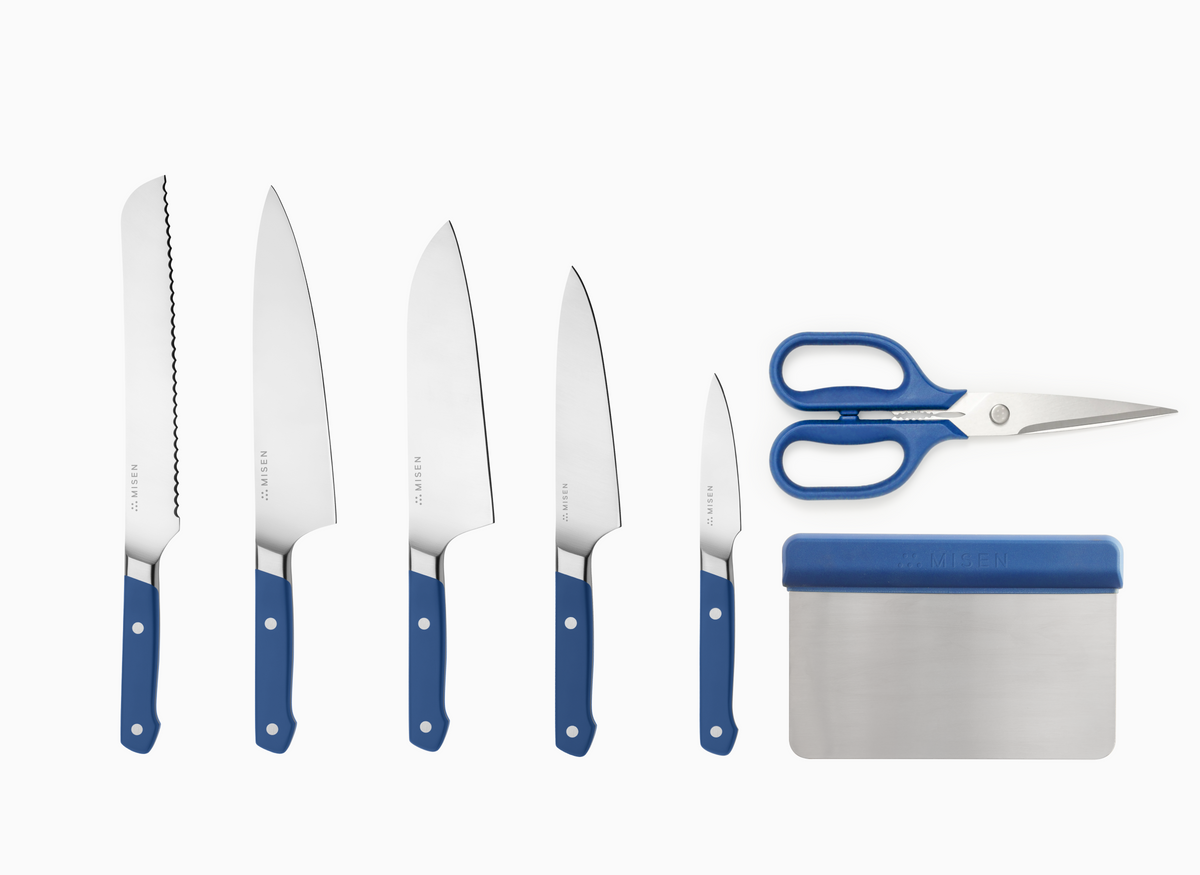 A 7 pc knife set with blue handles and AUS-10 steel blades, arranged vertically against a white background. The leftmost knife has an 8" serrated blade. The 2nd knife is an 8" chef's knife. The 3rd knife is an 8" santoku knife. The 4th knife is a 5.5" utility knife. The 5th knife is a 3.5" paring knife. To the right of the knives, there is a pair of standard kitchen shears and a rectangular bench scraper, both with blue handles. Each item has the word "MISEN" visible.