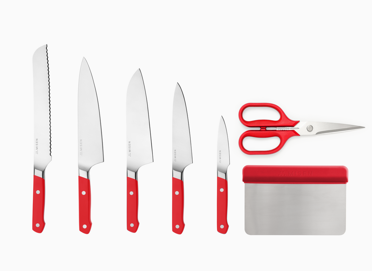 The Misen 7-piece Knife Set in red comes with an 8 inch Serrated Knife, an 8 inch Chef's Knife, an 8 inch Santoku Knife, a 5.5 inch Utility Knife, a 3.5 inch Paring Knife, a pair of Standard Kitchen Shears and a Bench Scraper.