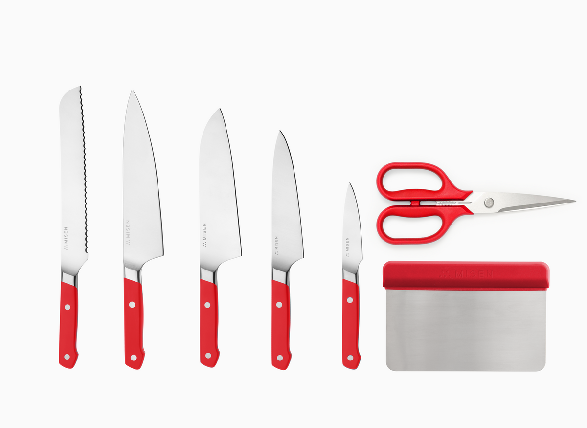 A 7 pc knife set with red handles and AUS-10 steel blades, arranged vertically against a white background. The leftmost knife has an 8" serrated blade. The 2nd knife is an 8" chef's knife. The 3rd knife is an 8" santoku knife. The 4th knife is a 5.5" utility knife. The 5th knife is a 3.5" paring knife. To the right of the knives, there is a pair of standard kitchen shears and a rectangular bench scraper, both with red handles. Each item has the word "MISEN" visible.