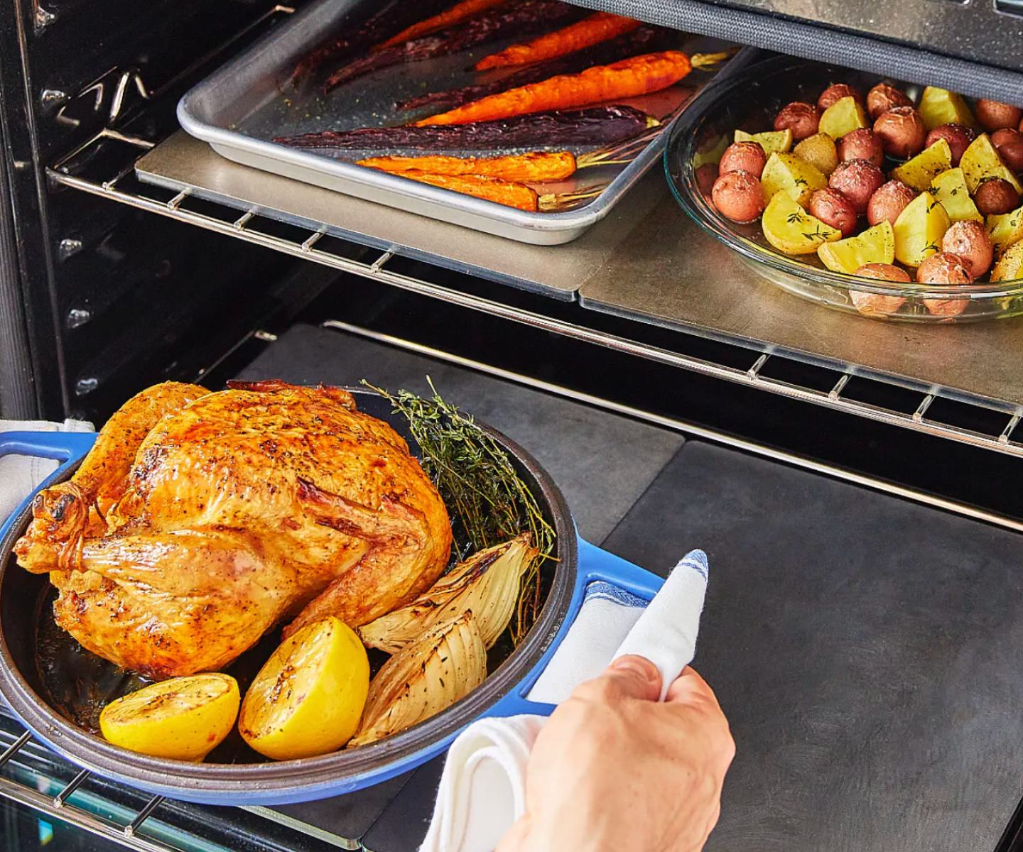 Whole roasted chicken with lemon and onion in blue pan being placed in oven alongside roasted carrots and potatoes
