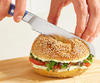 Using a short 6.5 inch blue serrated knife to cut a sesame bagel sandwich containing sprouts, tomato, and cream cheese in half.