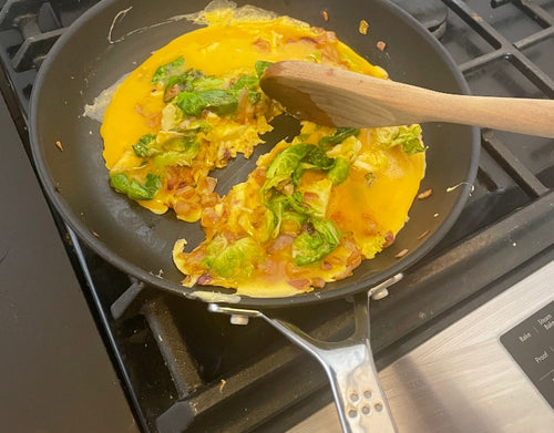 A top-view of scrambled eggs with greens and onions being made in a Misen Nonstick Pan. An unseen hand uses a wooden spoon to stir the eggs.