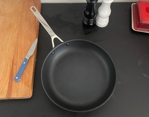 A bird’s eye view of a Misen Nonstick Pan on a black countertop. To the left of the pan is a blue Misen Paring Knife.