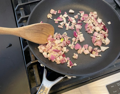 An unseen hand uses a wooden spoon to stir chopped red onion in a Misen Nonstick Pan, which sits on a stovetop.