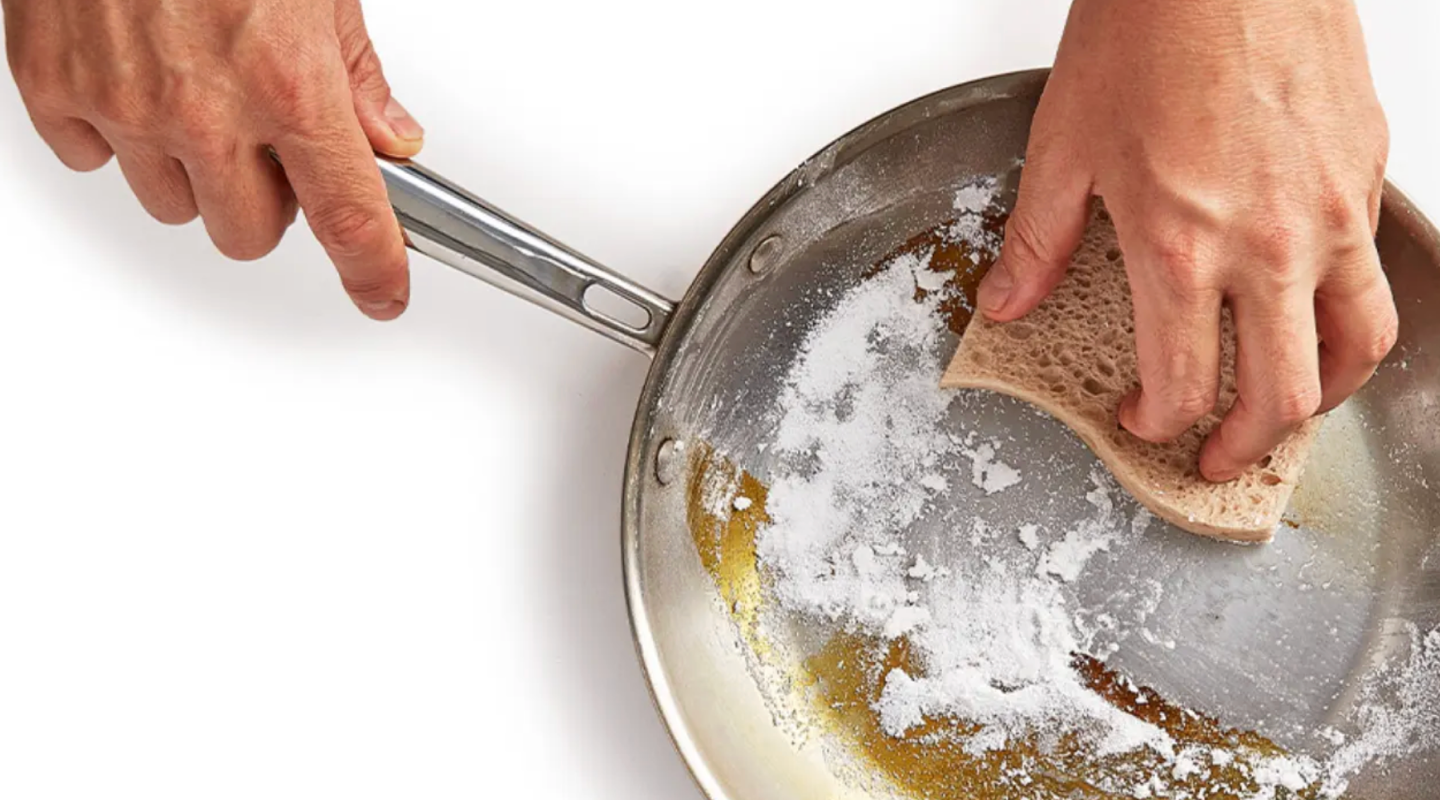 Cleaning a stainless steel pan with baking soda and a sponge to remove burnt residue
