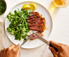 An overhead shot of a person cutting a sliced steak with a Misen straight-edged steak knife on a white plate. The steak is served alongside a pile of arugula salad dressed with olive oil and salt, and two lemon wedges. The person is holding a fork in their left hand and a knife with a black handle in their right hand, with the word "MISEN" visible on the blade. A glass of what appears to be beer is partially visible in the upper right corner.