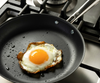 A sunny-side up egg cooking in a Misen Carbon Nonstick™ Frying Pan on a gas stove. The egg white is crispy around the edges, and small droplets of oil are visible in the pan.