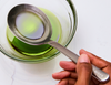 A close-up view shows a stainless steel Misen Measuring Chef Spoon resting in a clear glass bowl containing a green olive oil. The spoon is etched with measurements for 1 tbsp and 1 tsp.