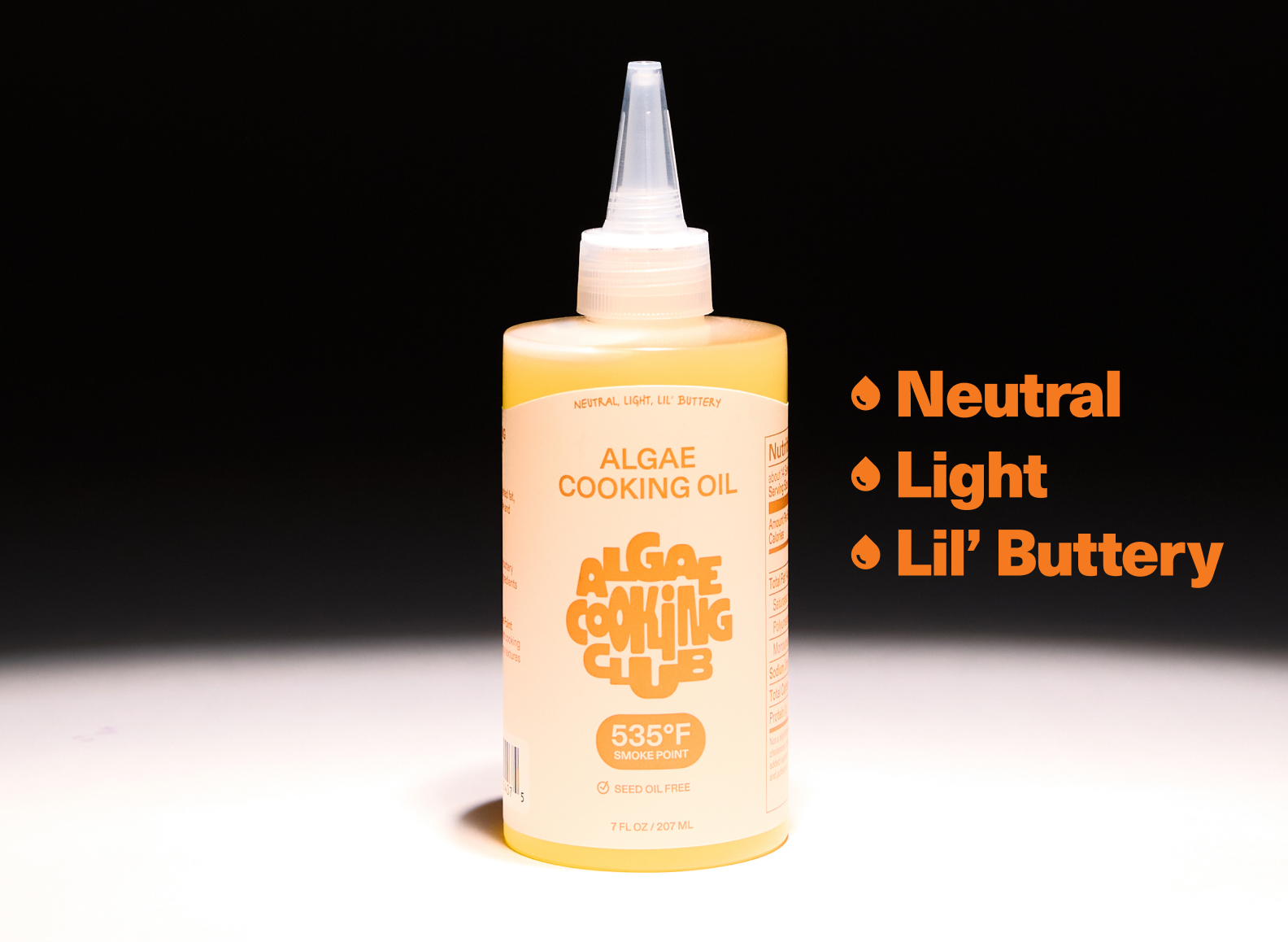 A pale yellow bottle of Algae Cooking Club cooking oil is centered against a dark background. The bottle has a tapered, clear plastic nozzle top. The label on the bottle is a lighter shade of yellow with orange text. The top of the label reads "NEUTRAL LIGHT, LIL' BUTTERY" in small, capitalized letters. Below this, in larger, capitalized letters, it says "ALGAE COOKING OIL" and "ALGAE COOKING CLUB." Underneath the brand name, a circular graphic indicates a "535°F SMOKE POINT" and "SEED OIL FREE."