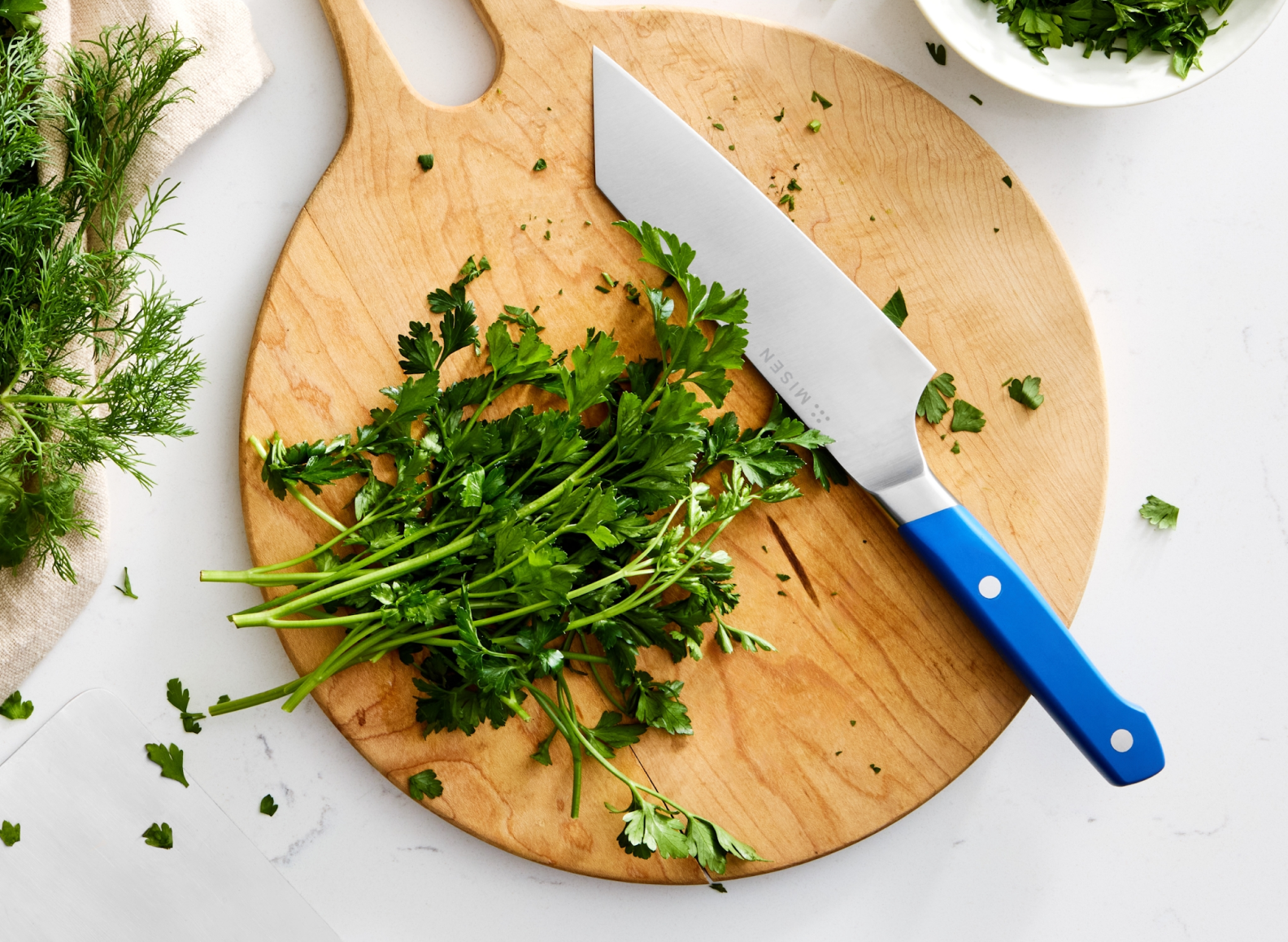 A top-down shot shows a large pile of fresh parsley on a round wooden cutting board. A 6 inch ACUTO440 bunka knife with a blue handle, labeled "MISEN," rests on the board to the right of the parsley. To the left of the cutting board, a Misen kitchen towel holds a bunch of fresh dill. In the upper right corner, a white bowl contains more chopped green herbs, likely parsley.