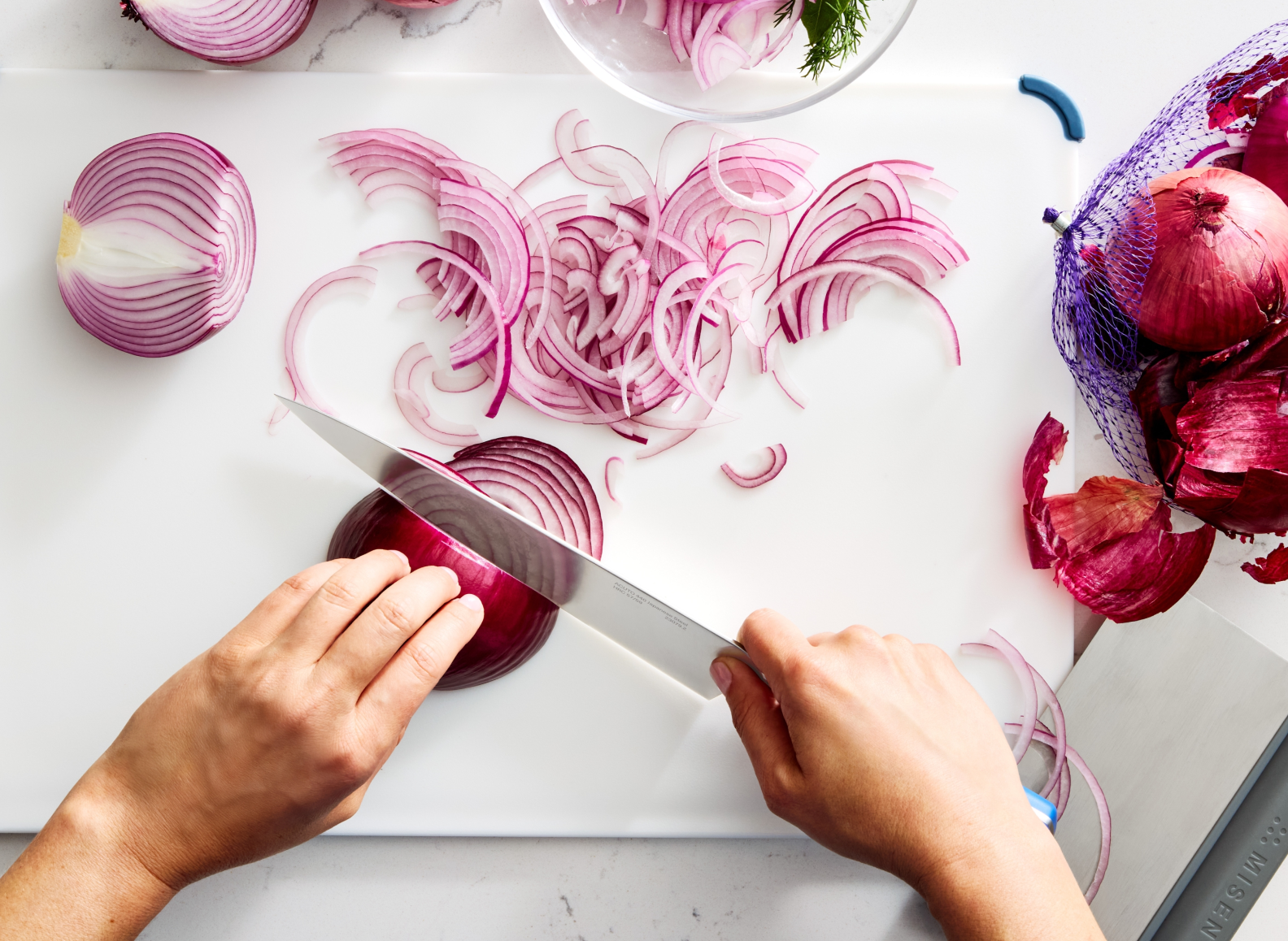 A top-down view shows a person slicing a red onion on a white cutting board with an ACUTO chef's knife. Many thin, purple onion slices are scattered across the cutting board. To the left, half of a red onion sits on the board. To the right, a purple mesh bag filled with red onions and onion skins rests next to the cutting board. In the upper right corner, a clear bowl contains more onion slices and dill. A gray bench scraper with the word "MISEN" embossed on the handle is visible in the bottom right corner.